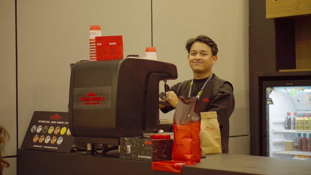 A barista at the counter beside the Tanamera branded machine, retail coffee bags and a drinks fridge behind