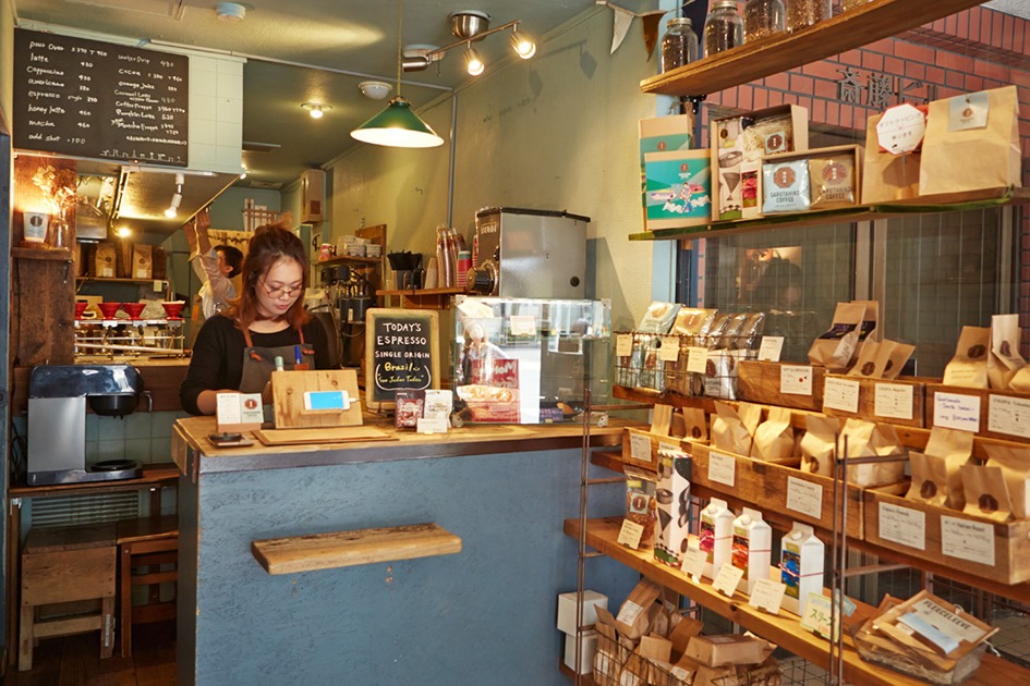 A barista working the counter at Sarutahiko Coffee's Ebisu flagship, shelves of branded retail bean bags lining the wall beside the register