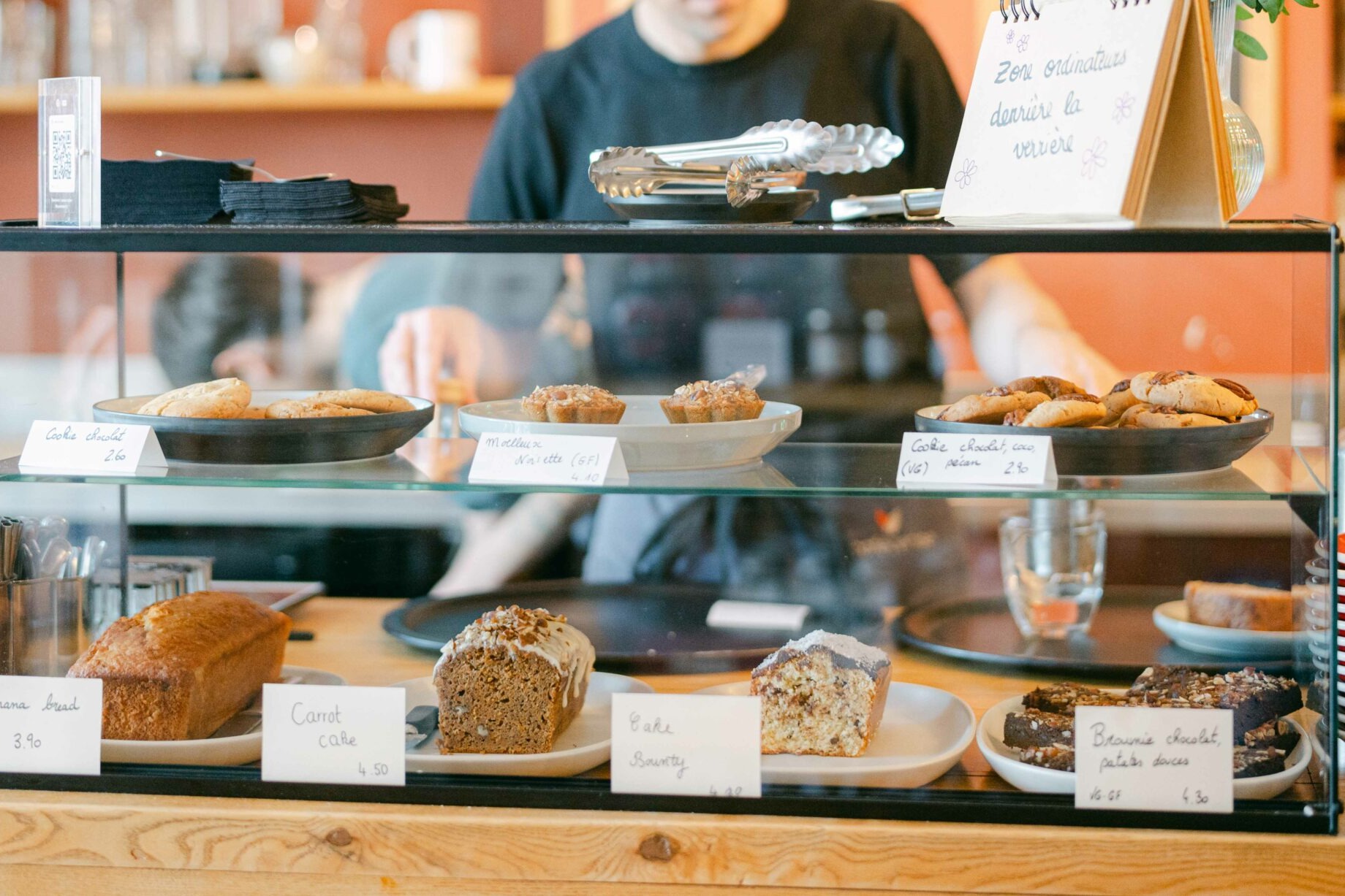 The glass pastry case at Le Cafe Cerise, banana bread, carrot cake and cookies labeled with handwritten price cards, a staff member at the counter behind