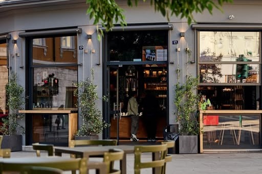The storefront entrance of Urban Caffè in the evening, terrace tables and potted plants out front, a customer entering the lit doorway