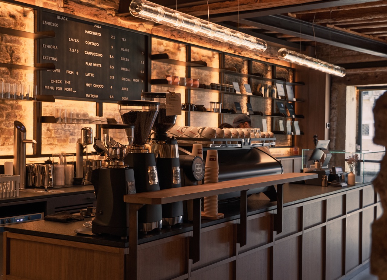 The wood and exposed brick counter at Caffè Cannaregio with a menu board, espresso machine, and grinders under a canal side window
