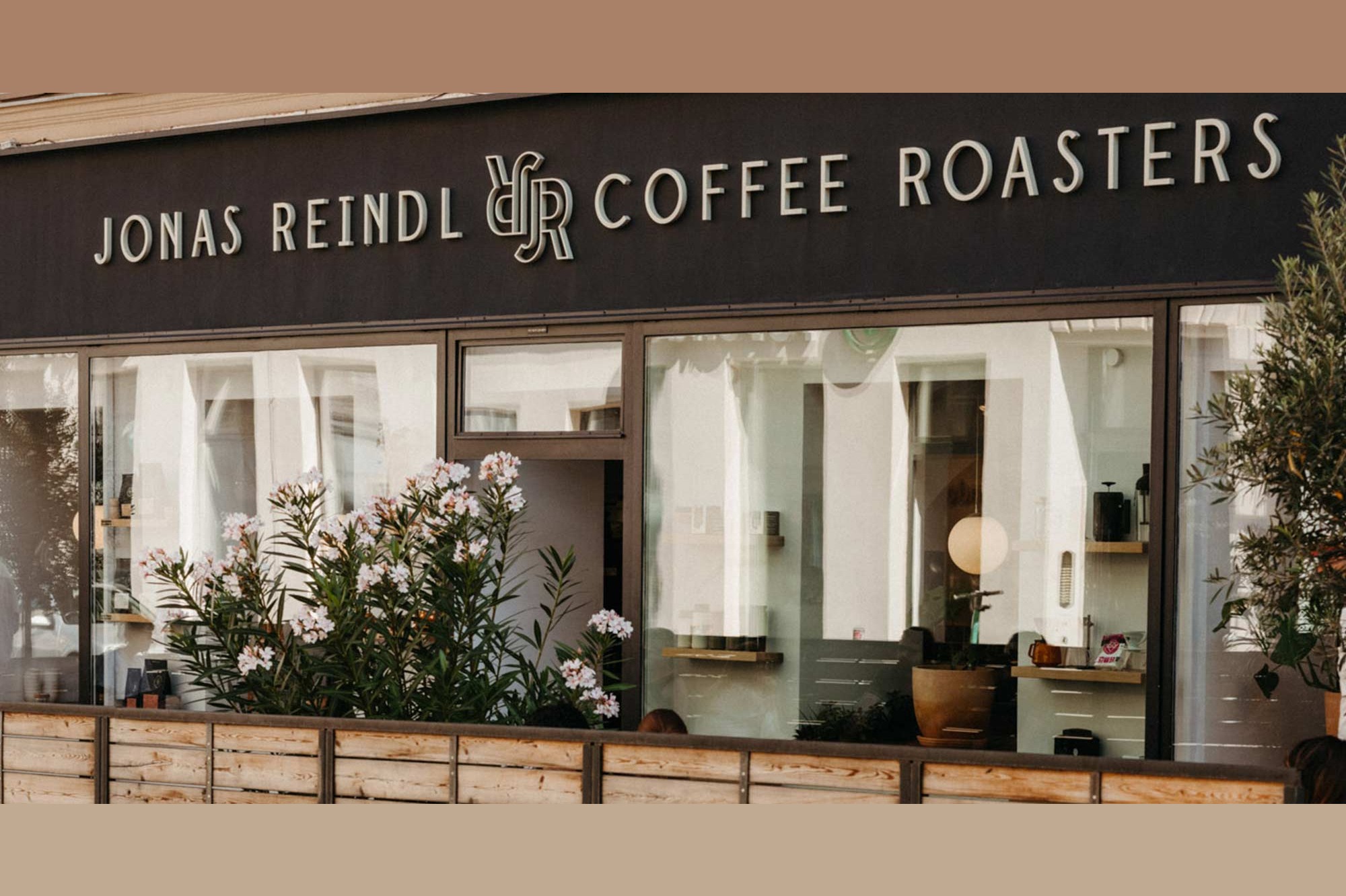 The storefront of Jonas Reindl Coffee Roasters, full signage above large windows, flowering oleander plants and roastery shelves visible inside